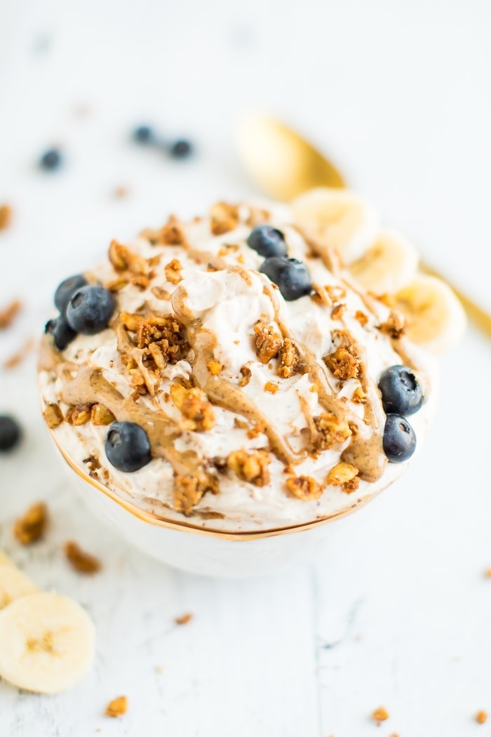 A bowl of yogurt covered with peanut butter drizzle, grain-free granola, blueberries and bananas in a gold rimmed bowl on a white wooden board. . A gold spoon in the background.
