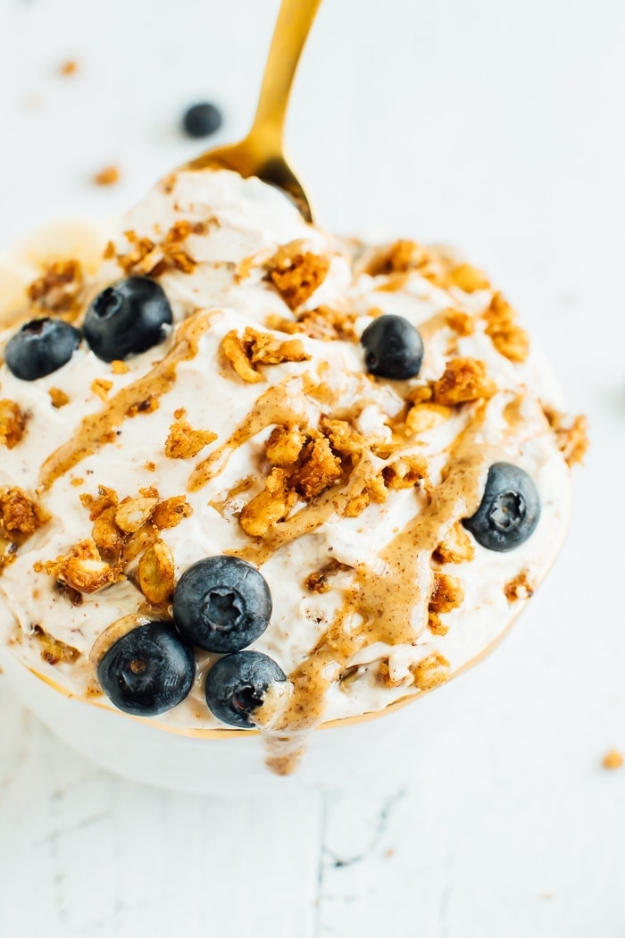 A delicious yogurt bowl made with peanut butter, grain-free granola, blueberries and bananas in a gold rimmed bowl with a gold spoon on a white wooden board. A bowl of yogurt covered with peanut butter drizzle, grain-free granola, blueberries and bananas in a gold rimmed bowl on a white wooden board. . A gold spoon is in the bowl.