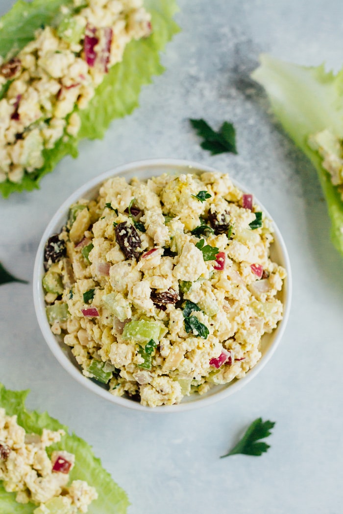 Overhead photo of Vegan Chicken Salad made with tofu, celery, almonds and raisins in a white bowl on white countertop; salad served in romaine.