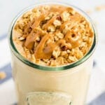 Cinnamon oatmeal cooke protein smoothie in a glass topped with granola and a peanut butter drizzle. Glass is on a striped napkin.
