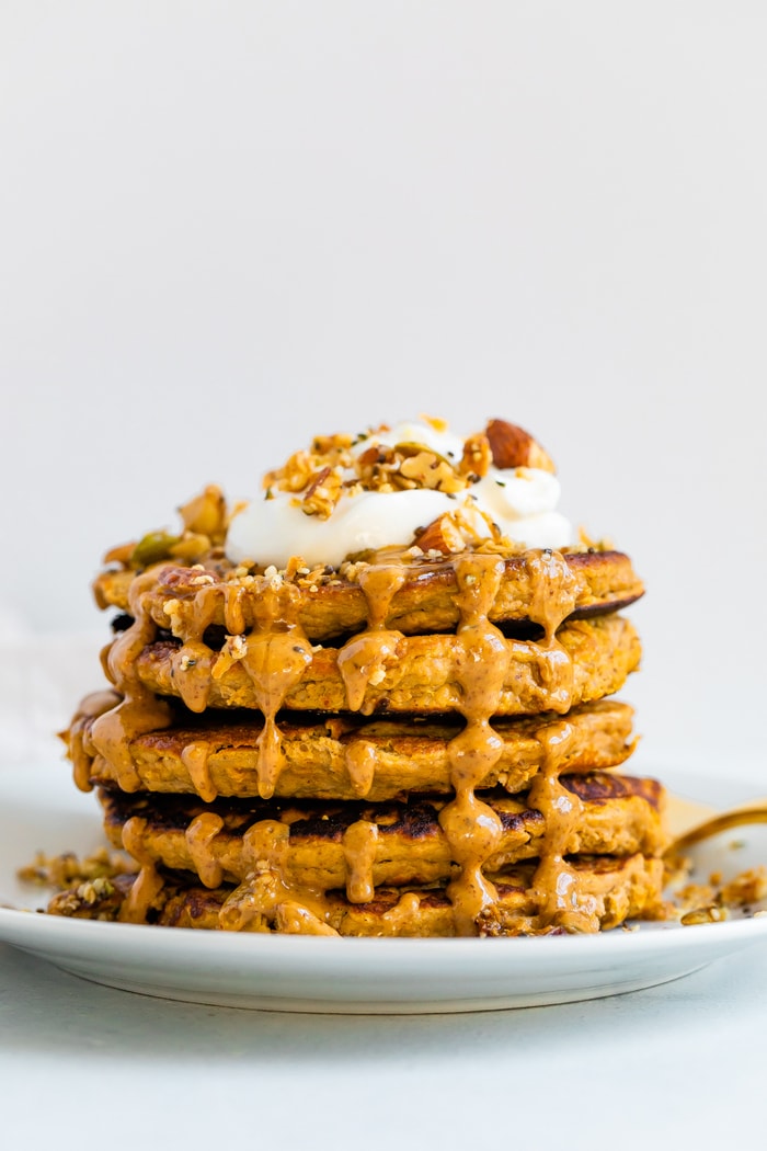 Stack of butternut squash pancakes topped with peanut butter, maple syrup, granola, and yogurt. A slice is taken out of the stack and a fork is resting on the plate.