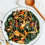 A large bowl of kale salad with sliced avocado on top. Wooden spoon is on the side of the bowl.