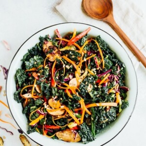 A large bowl of kale salad with sliced avocado on top. Wooden spoon is on the side of the bowl.