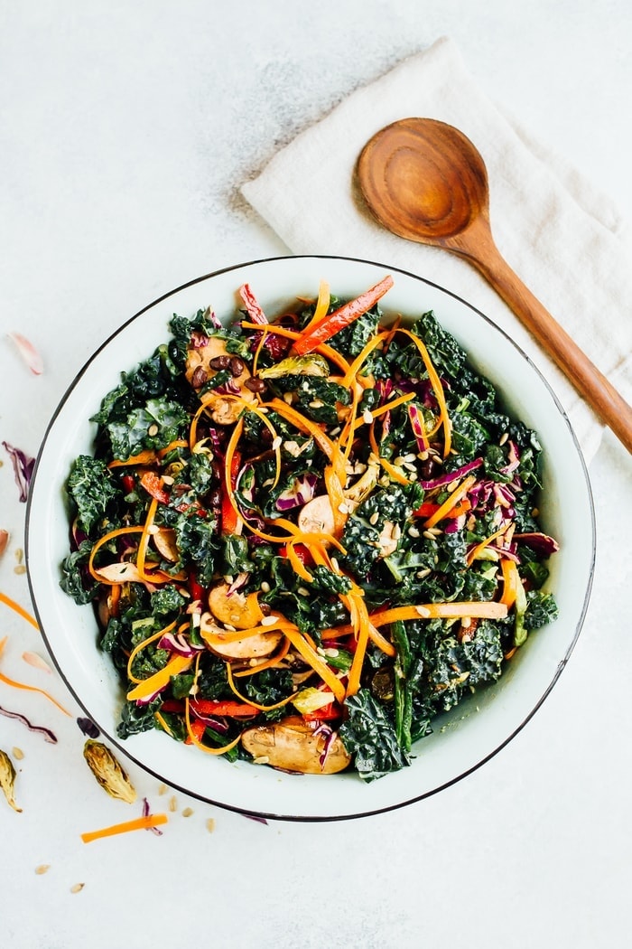 Pin this image to save this delicious detox kale salad recipe! A large bowl of kale salad with sliced avocado on top. Wooden spoon is on the side of the bowl.