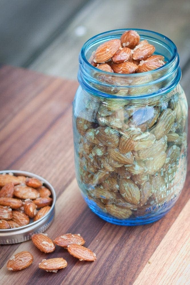 Sweet and delicious roasted almonds with maple syrup and coconut. Perfect for snacking! A blue mason jar filled to the top with maple coconut roasted almonds. The lid is laying next to the jar with additional almonds in it and a few scattered in front of it.