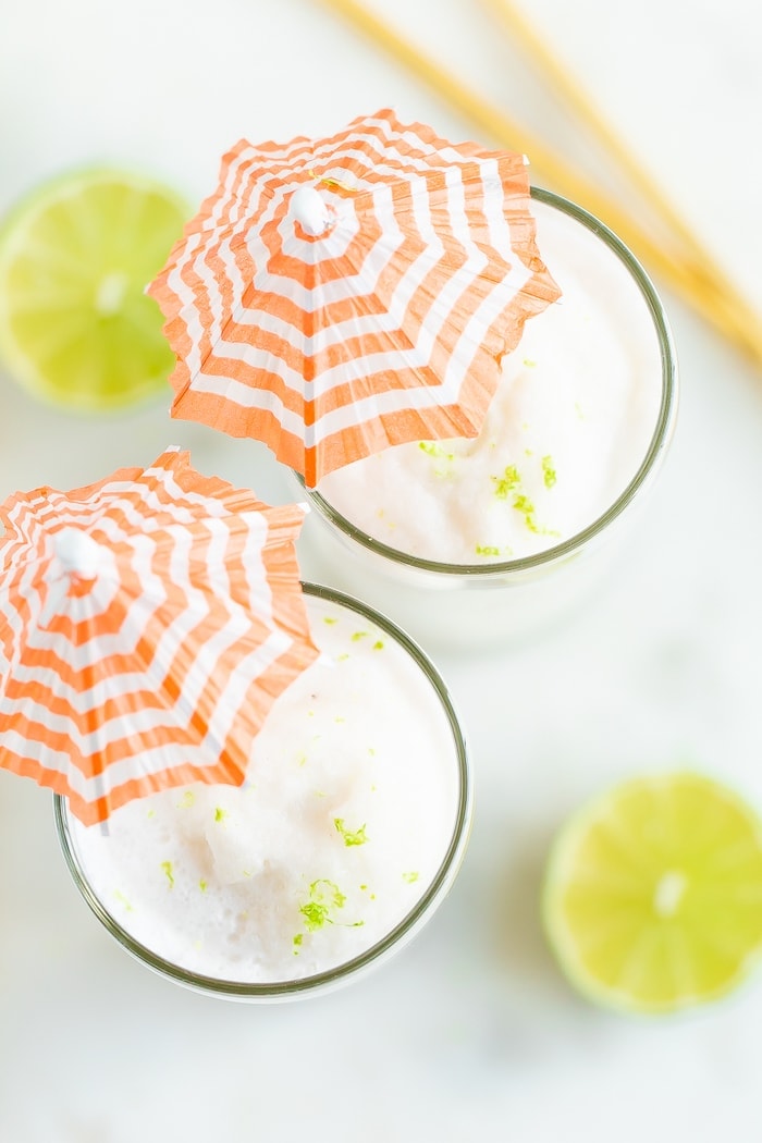 Overhead photo with two skinny coconut daiquiri cocktails in tall glasses with orange and white striped cocktail umbrellas.