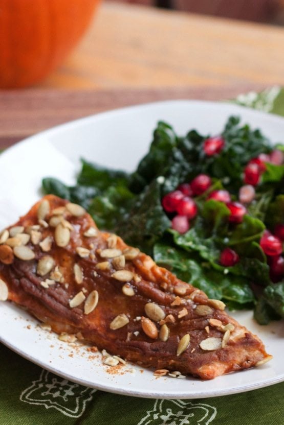 Pumpkin Spice Salmon A filet of pumpkin spice salmon on a white plate next to a kale salad with pomegranate arils.