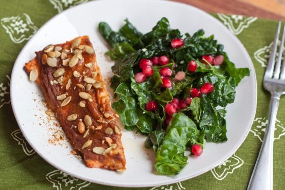 Pumpkin Spice Salmon A filet of pumpkin spice salmon on a white plate next to a kale salad with pomegranate arils. A fork is laying next to the plate.