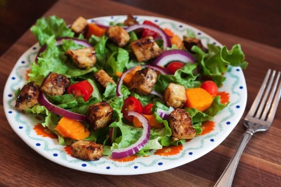 Maple Lemon Tempeh Cubes -- Great for Topping Salads or Snacking An overhead photo of a bed of salad greens with red onion slices and maple lemon tempeh cubes on a dinner plate. There is a silver fork laying next to the plate.