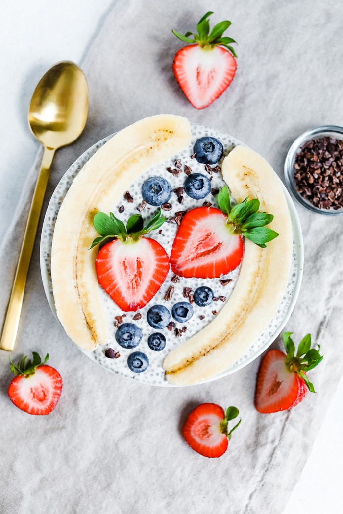 Bowl with chia pudding topped with a split banana, strawberries, blueberries and cacao nibs. A gold spoon, slices strawberries, and small bowl of cacao nibs are beside the bowl.