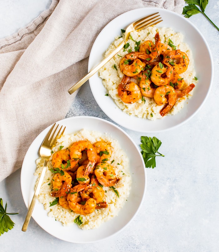 Two plates of low carb cauliflower cheese grits topped with blacked shrimp and fresh parsley. Two forks are resting on the plates, and a cloth napkin in on the table.