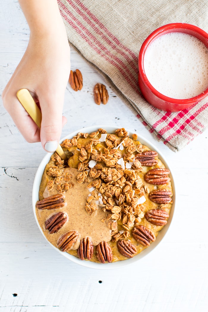 This protein pumpkin oatmeal recipe is loaded with pumpkin flavor and protein. The secret is whipping egg whites into the oats while they cook! Hand taking a spoonful fo protein pumpkin oatmeal. A mug of almond milk is to the side.