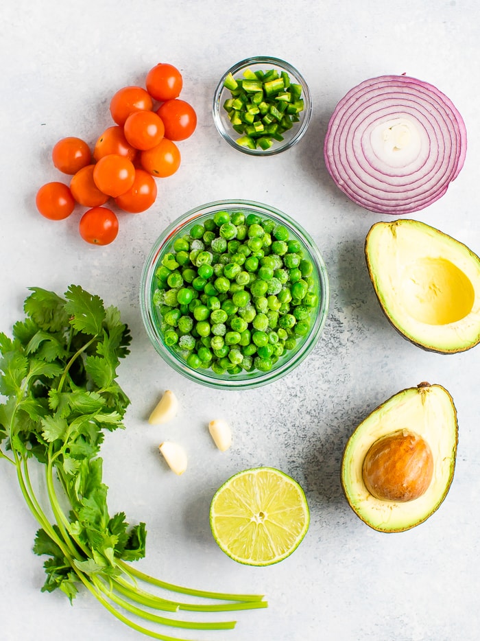 Ingredients for pea guacamole on a table: tomatoes, jalapeño, red onion, avocado, lime, garlic, cilantro and a bowl of frozen peas.