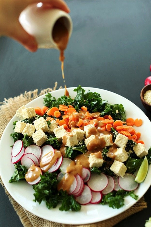Crunchy Thai Kale Salad by Minimalist Baker Crunchy Thai Kale Salad on a white plate. Hand in upper left corner pouring dressing over salad