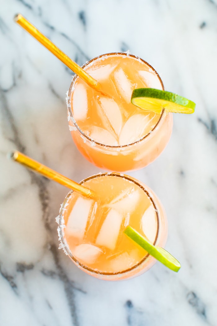 Two paloma drinks in glass cups with ice, lime wedges and gold straws.