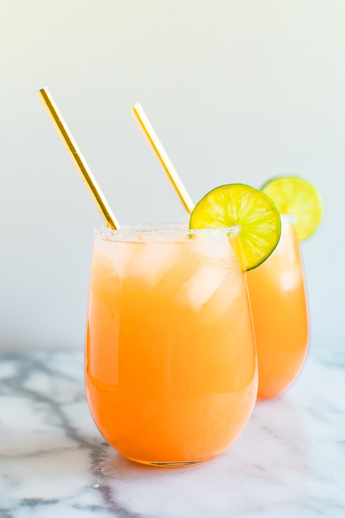 Two paloma drinks in glass cups with ice, lime wedges and straws on white marble table.