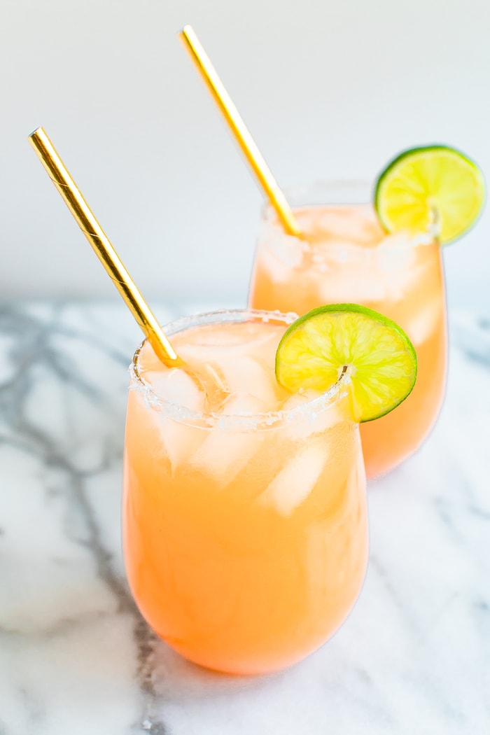 Two paloma drinks in stemless glasses with ice, lime wedges and gold straws.