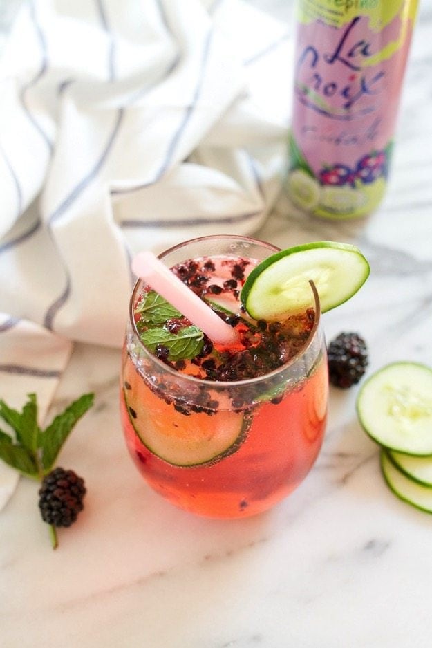 A stemless wineglass filled with blackberry cucumber mojito, topped with berries and cucumbers. There is a cucumber slice on the rim of the glass and a pink straw. There is a LaCroix can in the background.