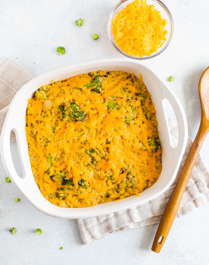 Broccoli quinoa casserole with cheddar cheese in a baking dish. Wooden spoon and glass bowl with cheddar cheese on the side.
