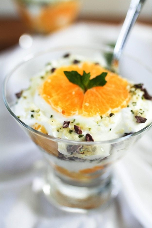 Clementine Greek Yogurt Parfaits with layers of creamy yogurt and juicy clementines along with a Hemp Heart and cacao nib crumble Close up of a clementine slice on top of the greek yogurt parfait in a glass bowl.