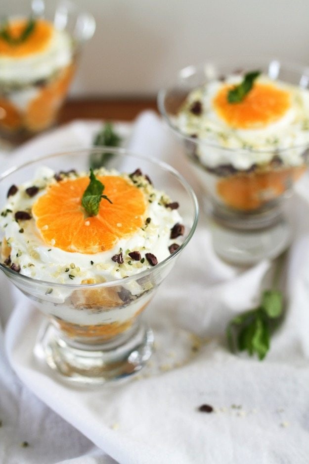 Clementine Greek Yogurt Parfaits with layers of creamy yogurt and juicy clementines along with a Hemp Heart and cacao nib crumble A glass bowl of clementine greek yogurt parfait on a white table cloth.
