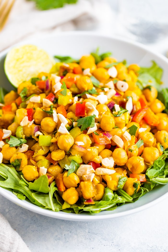 A bowl of curried chickpea salad made with chopped red onion, red pepper, cilantro, and dates, served over a bed of greens with a half of a lime to the side.