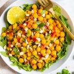 A serving of curried chickpea salad made with chopped red onion, red pepper, cilantro, and dates, served over a bed of greens. Half a lime and gold fork also in the bowl.