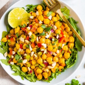 A serving of curried chickpea salad made with chopped red onion, red pepper, cilantro, and dates, served over a bed of greens. Half a lime and gold fork also in the bowl.