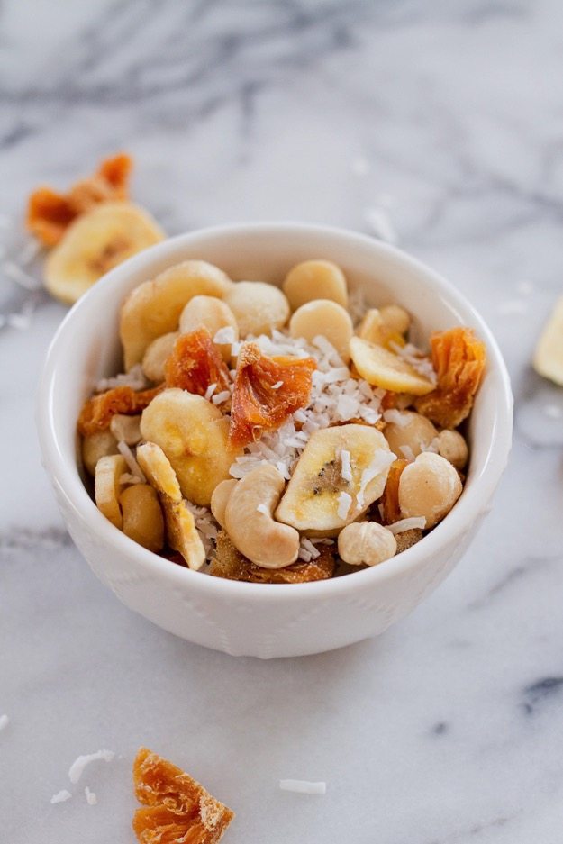 Take a trip to the tropics with this island-inspired tropical trail mix with cashews, macadamia nuts, banana chips, dried pineapple and coconut. Tropical trail mix served in a small white bowl.