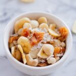 Tropical trail mix with cashews, macadamia nuts, banana chips, dried pineapple and coconut served in a small white bowl.
