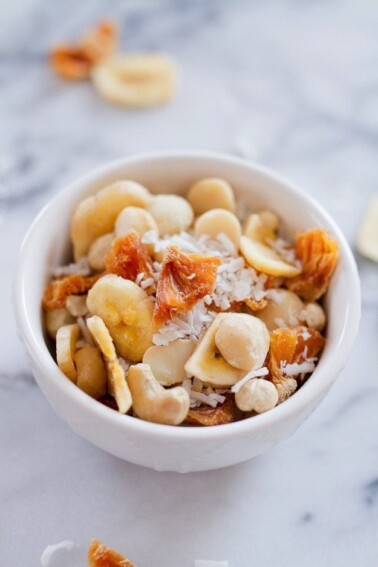 Tropical trail mix with cashews, macadamia nuts, banana chips, dried pineapple and coconut served in a small white bowl.