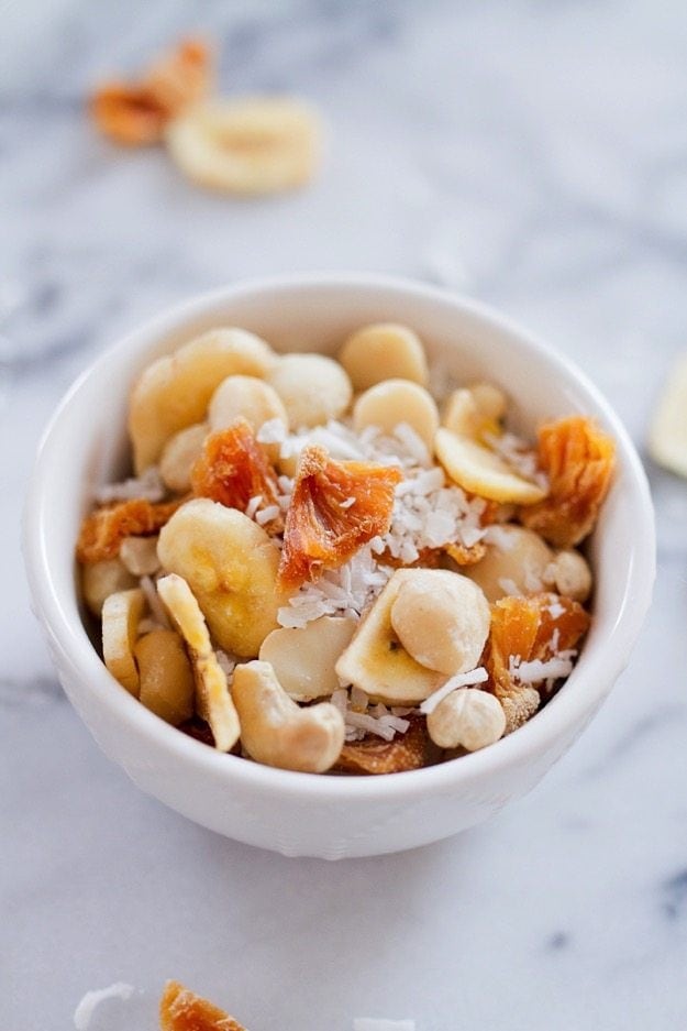 Take a trip to the tropics with this island-inspired tropical trail mix with cashews, macadamia nuts, banana chips, dried pineapple and coconut. Tropical trail mix with cashews, macadamia nuts, banana chips, dried pineapple and coconut served in a small white bowl.