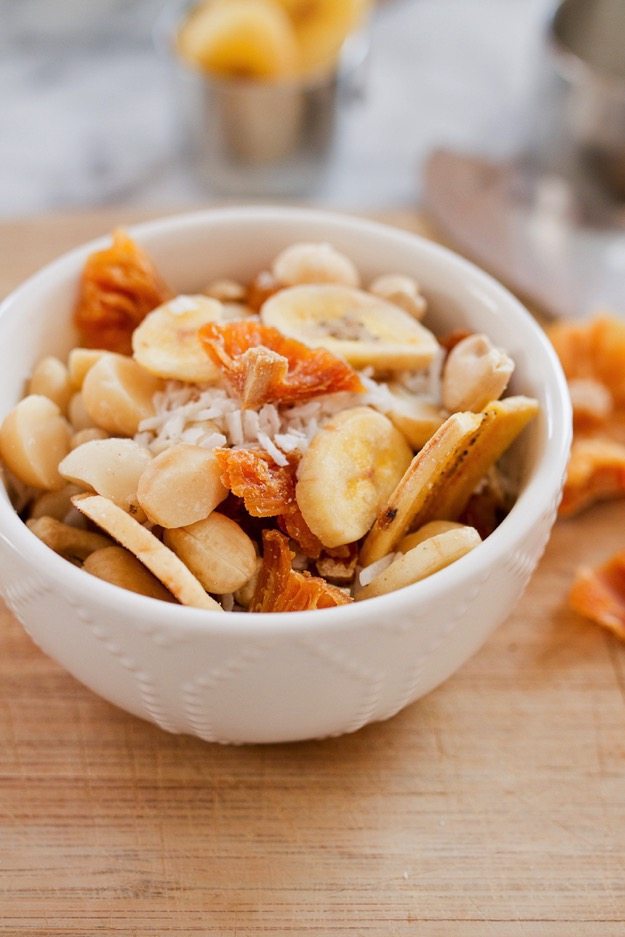 Take a trip to the tropics with this island-inspired tropical trail mix with cashews, macadamia nuts, banana chips, dried pineapple and coconut. Tropical trail mix in a small white bowl with cashews, macadamia nuts, banana chips, dried pineapple and coconut.