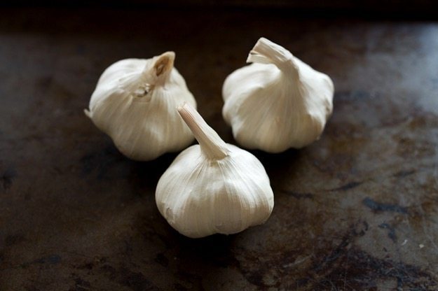 Pin this image to save a super simple recipe for how to make roasted garlic. Three heads of garlic sitting on a baking sheet.