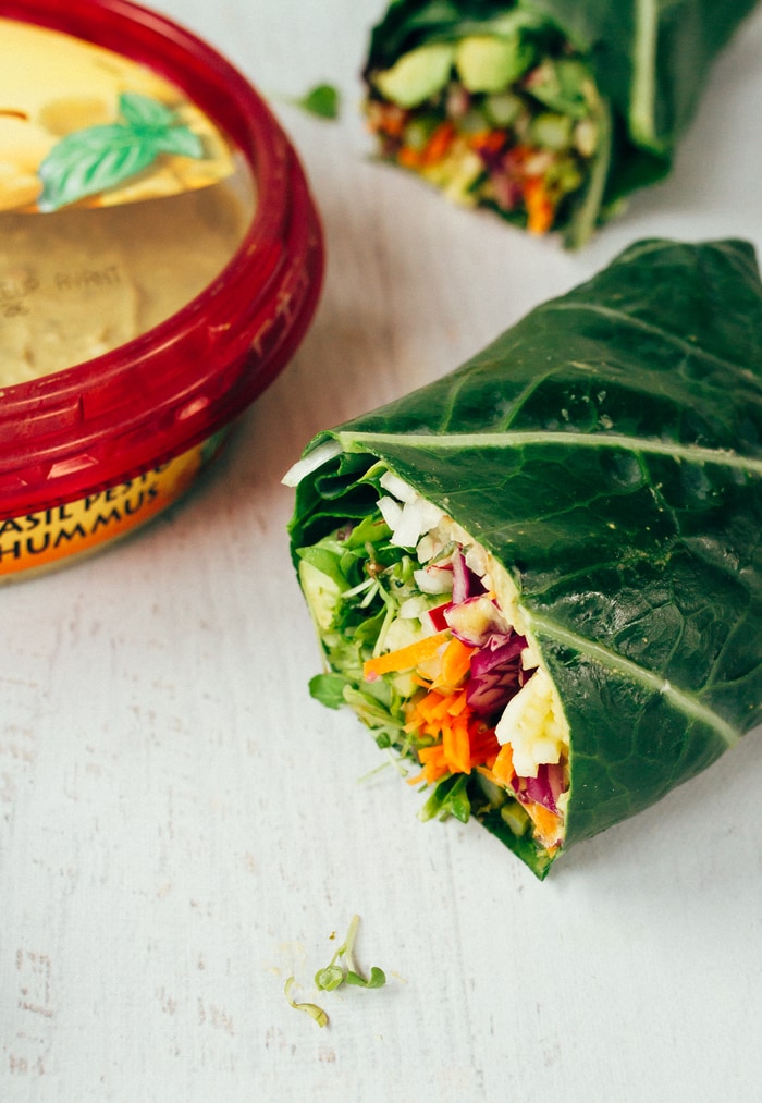 Save the recipe for these veggie packed collard wraps for later! Two halves of veggie packed collard green wraps sitting on a white countertop. Package of hummus on the edge of the picture.