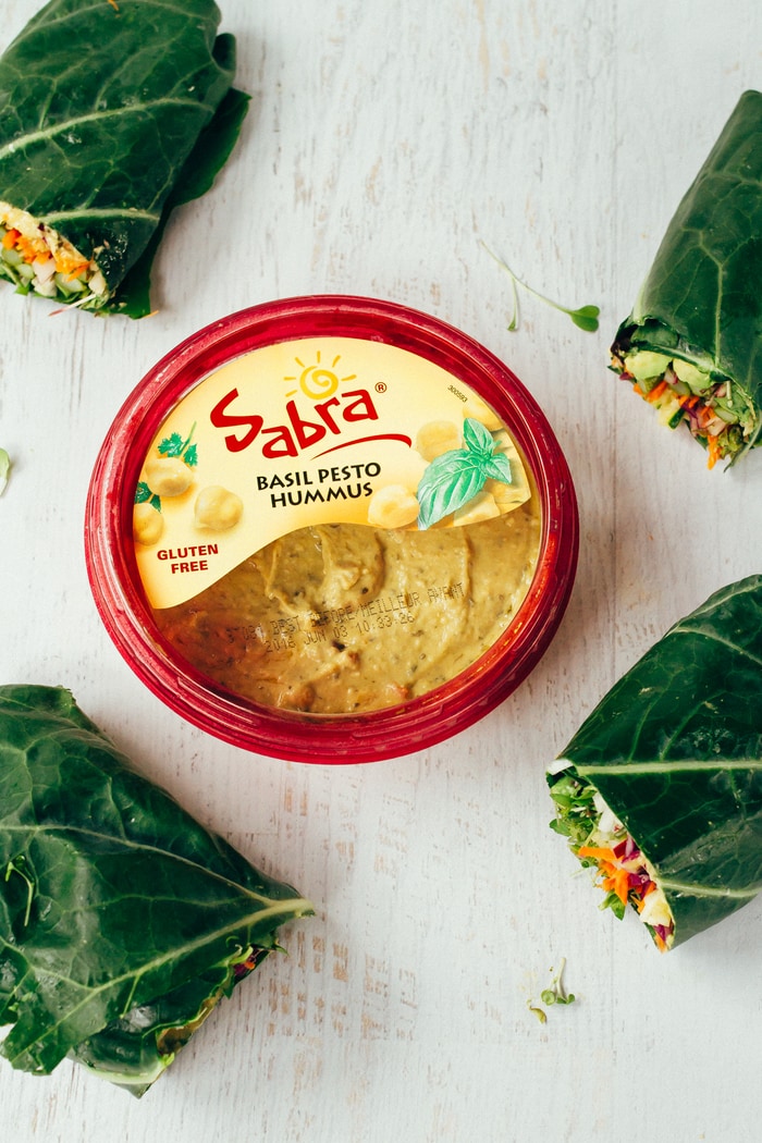 Pin this image to save these veggie packed hummus collard green wraps! Sabra basil pesto hummus container sitting on a white wood counter in the center of the frame. Four collard green wraps around the edge.