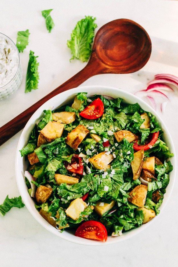Mustard Greens salad with roasted potatoes and tomatoes served in a white bowl next to a wooden spoon. 