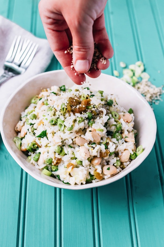 This Mediterranean-inspired rice salad with creamy cannellini beans and Hemp Hearts can be served warm or chilled and makes a lovely vegetarian main or a flavorful side. A woman\'s hand sprinkling hemp seeds on top of the lemony rice salad in a white bowl.