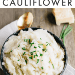 Bowl of mashed cauliflower topped with chives. Serving spoon in bowl. Bowl is on a napkin and wood table with cheese and chives to the side. Text above says "6-ingredient healthy mashed cauliflower".