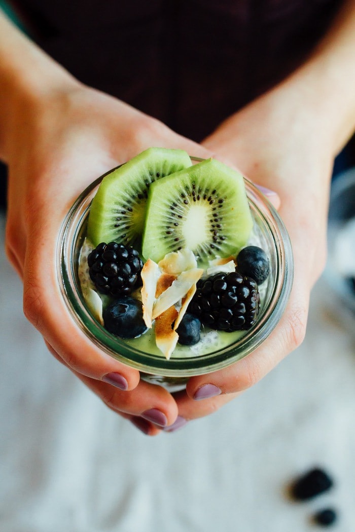 This Kiwi Chia Pudding Parfait is a unique way to enjoy fresh kiwi. The combo of chia seed pudding and kiwi puree makes for a healthy breakfast or snack that’s filling and delicious. Woman\'s hand holding a kiwi chili pudding jar.