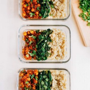 Three curried chickpea bowls served in glass tupperware containers alongside brown rice and spinach.