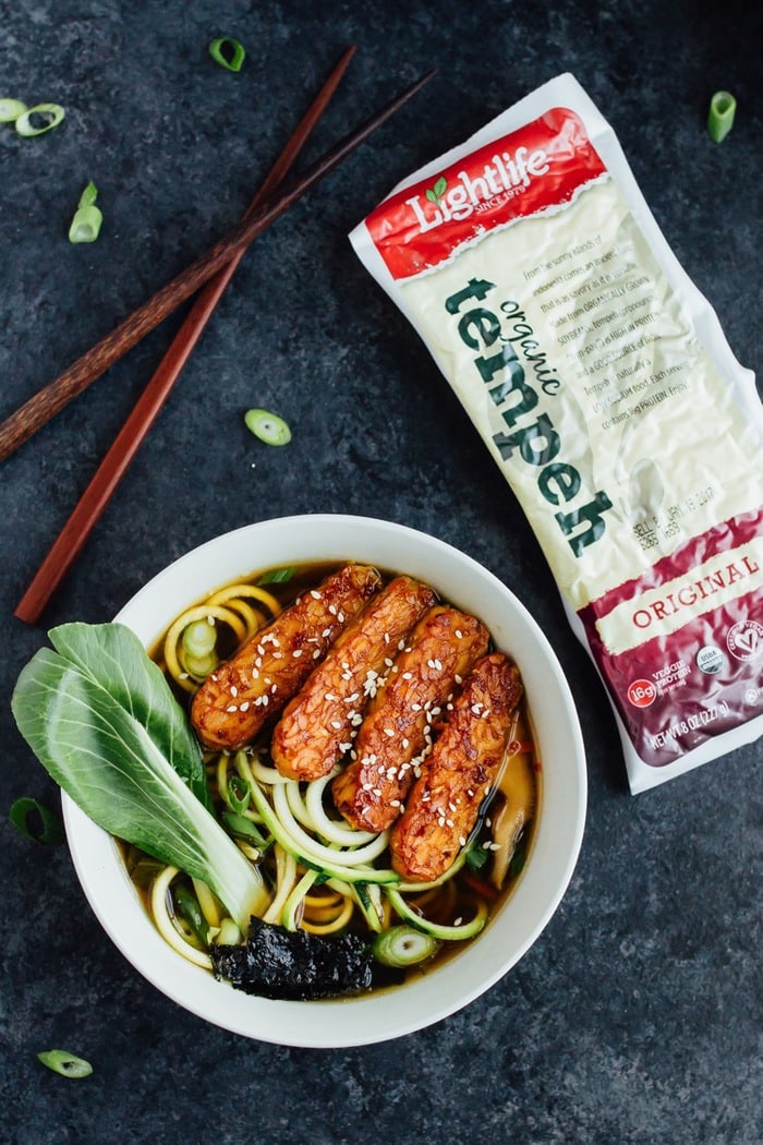 Overhead photo of vegan zucchini noodle ramen bowls served in a white bowl with wood chopsticks on dark grey countertop with Lightlife Tempeh.