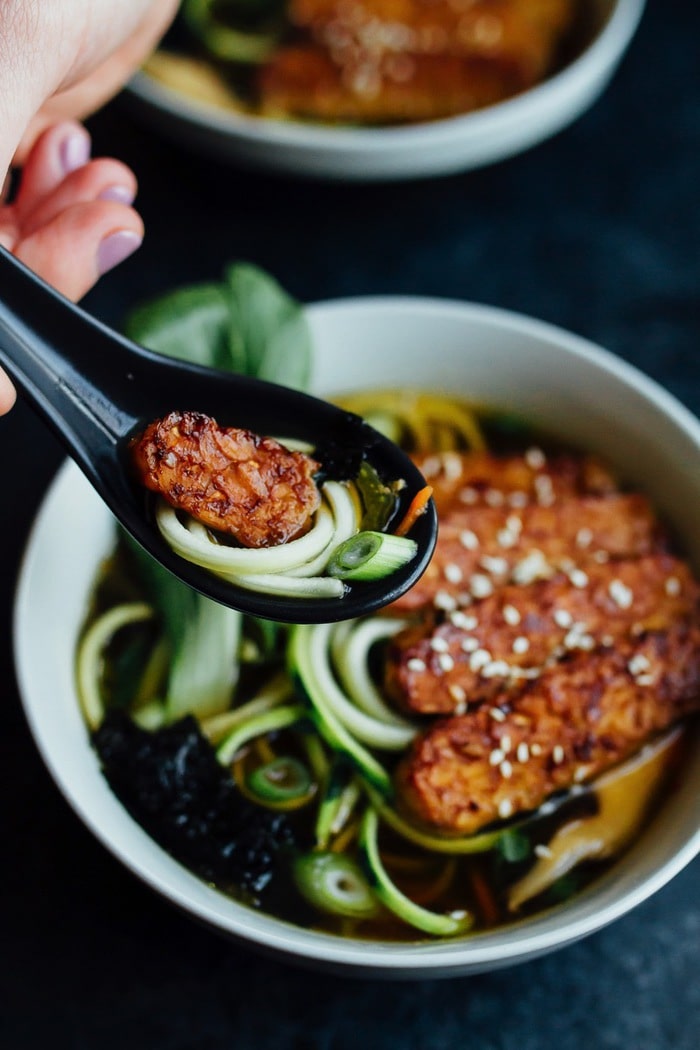Hand holding black spoon of tempeh and zucchini noodles, over a bowl of vegan zucchini ramen.