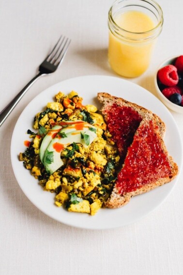 Tofu scramble topped with avocado slices and a drizzle of hot sauce next to two slices of toast topped with fruit jam.