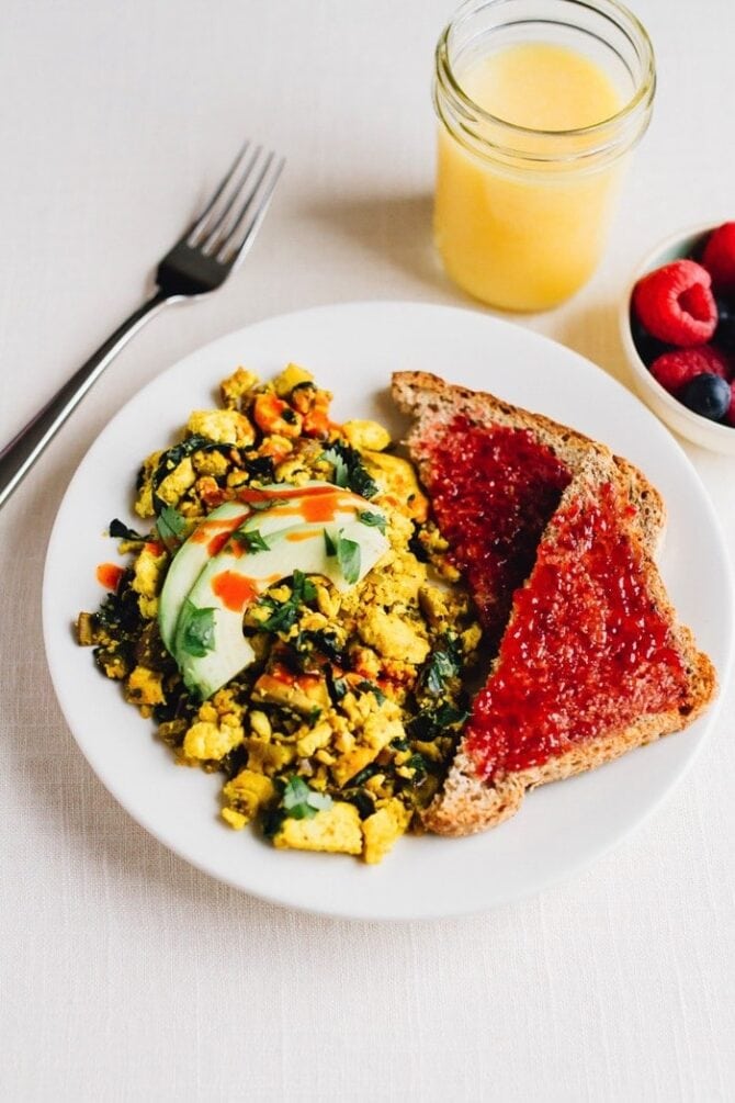 golden tofu scramble on a plate next to two pieces of toast with jam. A fork, bowl of berries and a glass of orange juice are next to the plate.