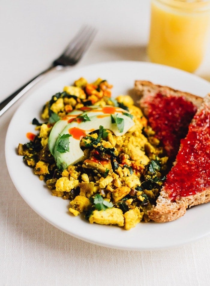 Turmeric tofu scramble on a plate with avocado on top. Two slices of bread with jam on the side. Glass of orange juice and fork also on the side.