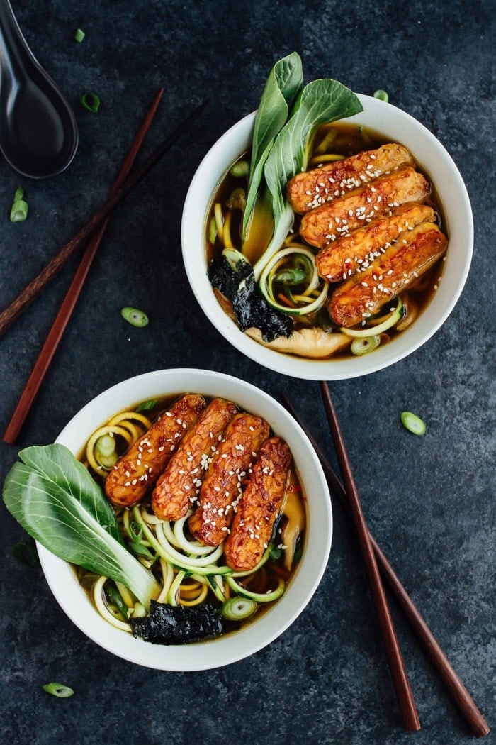 Overhead photo of vegan zucchini noodle ramen bowls served in white bowls with wood chopsticks on dark grey countertop.