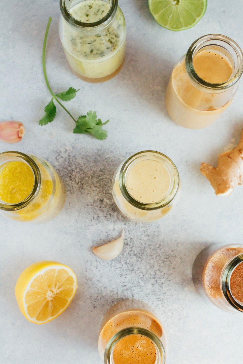 Healthy homemade salad dressings in a glass jars spread across a counter top with fresh garlic, lime, ginger and cilantro in the background.