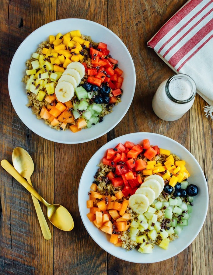 Overhead photo of Cinnamon Quinoa Breakfast Bowls with chopped fresh fruit, served in white bowls on wood table.