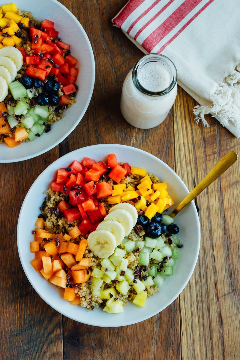 Overhead photo of Cinnamon Quinoa Breakfast Bowls with chopped fresh fruit, served in white bowls on wood table.